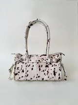 Cartera de cuero con textura de pelo vacuno y estampado animal print tipo dálmata. Presenta una estructura rígida, doble asa de hombro, cierre superior y detalles decorativos de tiras con tachas, hebillas metálicas y lazos laterales.