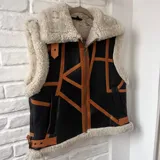 Chaleco de cuero negro y apliques de cuero marrón tostado, con interior y detalles de corderito sintético blanco crudo, cierre frontal y bolsillos.