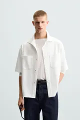 Sobrecamisa negra de tejido con elasticidad, cuello solapa y manga corta. Tiene dos bolsillos de plastrón en el pecho y cierre frontal con botones a presión.