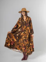 Vestido largo de manga larga, corte oversized y cuello camisero con abertura en V y botones. Presenta un estampado botánico en tonos tierra sobre fondo marrón oscuro, con volados en el ruedo.
