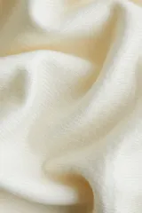 Buzo corto de punto fino en color crema, con diseño de un solo hombro y escote asimétrico. Presenta mangas largas y puños acanalados.