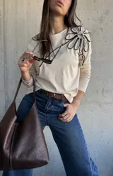 Remera manga larga de color beige, con detalle de aplique floral tridimensional en el hombro confeccionado con cordones en blanco y negro.