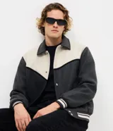 Campera de estilo varsity confeccionada en polyvelour, con diseño bicolor en gris oscuro y crema. Presenta cuello tipo polo, cierre frontal con botones a presión, mangas largas y puños y cintura con terminación en ribana a rayas.