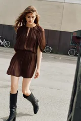 Vestido corto de tejido satinado color marrón, con cuello redondo, mangas largas con diseño fluido y cintura elástica. Presenta una abertura frontal y cierre posterior con botón.