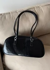 Cartera de hombro tipo baguette en color negro con acabado brillante efecto cuero arrugado. Presenta una estructura rectangular con bordes redondeados, cierre superior y doble asa fija.