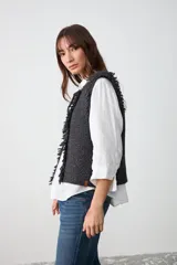 Chaleco de tejido de punto grueso en color negro, con diseño sin mangas, textura irregular y detalle de flecos en los bordes frontales. Cuenta con un cierre de lazo en la parte superior del cuello.