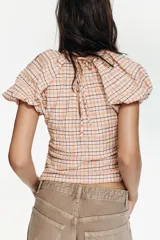 Blusa de manga corta abullonada con cuello redondo, confeccionada en tela a cuadros. Presenta detalle de frunces en los laterales y cierre lateral con cremallera oculta.