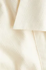 Camisa cropped de manga larga color crema, con cuello abierto y botones frontales. Presenta una textura sutil de tejido dobby y un calce ajustado con pinzas en la espalda.