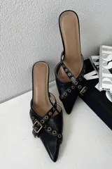 Mules de cuero negro con punta fina, diseño abierto en los laterales y tiras decorativas con ojales metálicos y hebilla ajustable.