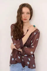 Blusa marrón semitransparente con estampado de lunares celestes, mangas largas abuchonadas, cuello en V con lazos y detalles de volados frontales.