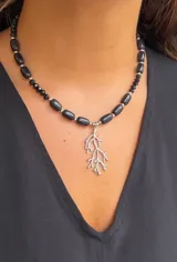 Collar corto de cuentas negras, algunas cilíndricas y otras facetadas, separadas por cuentas metálicas plateadas. Incluye un dije colgante plateado con forma de rama o coral.