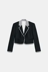 Blazer corta de color negro con cuello solapa, manga larga y hombreras. Presenta detalles combinados con estampado de cuadro vichy en el cuello, los bordes de la solapa, el ruedo y los puños. Cuenta con cierre frontal de un botón y falsos bolsillos delanteros de vivo.