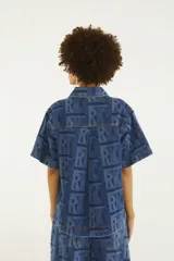 Camisa de jean de manga corta con calce recto, cuello clásico, bolsillos frontales con solapa y cierre de botones metálicos. Presenta un estampado gráfico con monograma en tonos de azul.