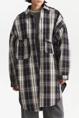 Camisa de largo extendido, estilo camisaco, con patrón de cuadros escoceses en tonos negro, blanco crema y gris lavanda. Presenta cuello camisero, cierre frontal con botones y dos bolsillos de parche con tapa en el pecho. El corte es holgado y la parte inferior es redondeada.