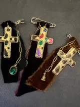Pin decorativo con forma de cruz dorada de estilo vintage, adornada con piedras incrustadas. La pieza incluye una base de tela de terciopelo desflecada, una cadena metálica y un pequeño dije colgante.