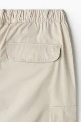 Pantalón cargo de algodón color beige con corte holgado. Presenta cintura elástica con cordón ajustable, múltiples bolsillos con solapa y botones a presión en las piernas y parte trasera, pliegues en las rodillas y cordones de ajuste en los bajos.