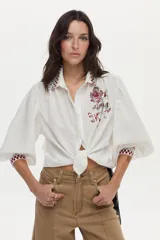 Blusa de mezcla de algodón y lino color blanco, con mangas abullonadas y cierre frontal de botones. Presenta detalles de bordado geométrico a cuadros en el cuello y los puños, y un gran bordado floral en el pecho.