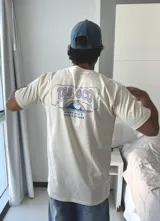 Remera de manga corta color beige jaspeado, con un estampado gráfico en la espalda que incluye el texto "TAS LOCO" y "SUNNY WITH A CHANCE OF STOKE" con un diseño de una persona en el agua con una ballena.