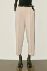 Pantalón tobillero de tiro medio, color beige, con pinzas delanteras y bolsillos frontales. Incluye un cinturón fino de cuero marrón con hebilla metálica dorada y cierre frontal con cremallera, botón interior y gancho.