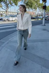 Blazer corto blanco de cuello solapa y manga larga con hombreras. Presenta un cierre frontal cruzado con dos botones dorados.