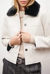 Chaqueta corta color beige con cuello de piel sintética negra, cierre frontal de botones y bolsillos laterales.