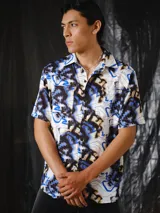 Camisa de manga corta de calce relajado, con cuello clásico, cierre frontal con botones y bolsillos aplicados en el pecho. Presenta un estampado integral abstracto en tonos crudo, negro, marrón y azul eléctrico.