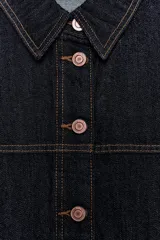 Cazadora de denim en color azul oscuro con costuras en contraste, cuello solapa, manga larga y cierre frontal con botones metálicos. Presenta dos bolsillos de plastrón en el delantero.