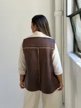Chaleco de cuero sintético color marrón chocolate con interior y solapas de corderito sintético color crudo. Diseño sin mangas con cierre frontal.