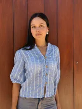 Camisa de corte recto y calce holgado, confeccionada en tela liviana color celeste con estampado de rayas verticales blancas con detalles de hojas. Presenta cuello en V, cierre frontal con botones de madera y mangas tres cuartos con terminación elástica.