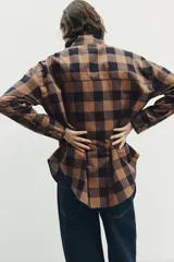 Camisa de manga larga con estampado de cuadros en tonos marrón y negro. Presenta un diseño cruzado con cuello vuelto y cierre frontal de botones, con un corte asimétrico en el bajo.