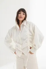 Camisa blanca de mangas largas con cuello clásico y cierre con botones. Presenta detalles de fruncido vertical en la parte delantera y cordones ajustables en los laterales. Corte holgado.