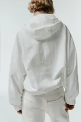 Chaqueta blanca de cuello redondo con capucha ajustable mediante cordones elásticos. Presenta manga larga con puños elásticos, bolsillos delanteros y bajo asimétrico ajustable con cordones y stoppers. El cierre frontal combina cremallera y botones ocultos bajo una solapa.