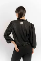 Blusa negra de lino con acabado brillante, de manga larga abullonada, escote con volados y cierre frontal con tiras para anudar.