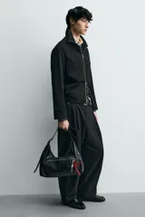 Bolso tipo bandolera de cuero sintético negro con acabado brillante. Presenta un compartimento principal con cierre de cremallera, un bolsillo frontal con cierre y detalle de charms colgantes decorativos. Incluye asa de hombro ajustable.