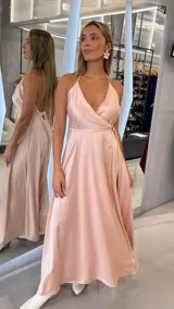 Vestido largo de satén color rosado, con diseño cruzado, escote en V pronunciado, tirantes finos tipo halter y lazo ajustable en la cintura.