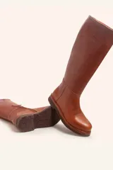 Bota de caña alta confeccionada en cuero vacuno suave, con diseño minimalista y suela de goma robusta. Presenta costuras visibles en la zona del empeine y un taco bajo cuadrado.