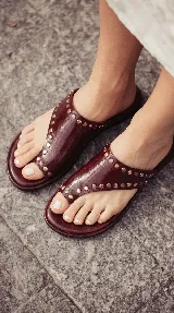 Sandalias chatas de cuero color bordó, con diseño de tira ancha que envuelve el empeine y sujeta el dedo gordo, decoradas con tachas metálicas doradas.