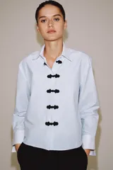 Camisa de algodón con cuello solapa y puños en color blanco a contraste. Presenta cierre frontal con alamares negros y bajo asimétrico redondeado.