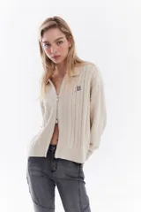 Sweater de punto color beige con cierre frontal de doble cursor, cuello polo y mangas largas con diseño de trenzas. Cuenta con un pequeño bordado en el pecho.