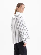 Camisa blanca de poplin con fit holgado, cuello con broche metálico y mangas largas con puños abotonados. Presenta un patrón de rayas verticales formadas por bordados de boquitas.