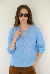 Blusa celeste de manga larga con delicados bordados florales en tono más claro en la zona del pecho y mangas. Presenta escote en V y puños elásticos abotonados.