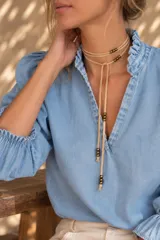 Gargantilla estilo corbatín confeccionada en tiento de cuero color beige, decorada con pequeñas cuentas metálicas doradas y extremos con flecos atados.