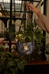 Cartera pequeña de cuero negro con textura tipo reptil, diseño semicircular y placa metálica dorada con logo en el frente. Cuenta con una correa ajustable y versátil con herrajes de bronce que permiten modificar su largo y forma.