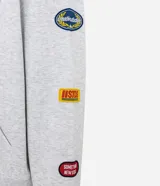 Buzo tipo hoodie con corte boxy, confeccionado en algodón con fondo melange. Presenta capucha, bolsillo canguro frontal, puños y ruedo acanalados, y múltiples parches bordados decorativos en el frente y las mangas.