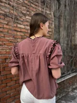 Blusa color chocolate con mangas cortas abullonadas, cuello redondo con volados y detalles de costura en zigzag de color rojo en el pecho y volados oscuros en los hombros.