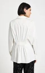 Camisa blanca de corte recto con cuello clásico estructurado, mangas largas con puños ajustados y cierre frontal de botones ocultos. Presenta un panel trasero con frunces y detalle elástico, con un bajo ligeramente redondeado.