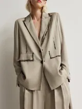 Blazer de corte recto confeccionado en viscosa, color gris topo. Presenta solapas clásicas, cierre frontal de dos botones y mangas largas con botones en los puños. Destaca por su diseño con detalles de efecto pintura en solapas, costuras y bolsillos. Incluye un bolsillo con solapa en el frente y abertura en la espalda.