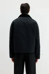 Campera negra de sarga encerada de algodón con cuello de pana. Presenta cierre frontal cubierto por una solapa con botones a presión, mangas raglán con puños ajustables mediante botones a presión, bolsillos insertados al bies y un bolsillo interior con botón a presión. Tiene un corte holgado y forro interior.