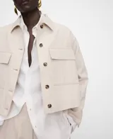 Chaqueta corta de algodón y lino color beige, de cuello italiano, manga larga con botones en los puños, cierre frontal de botones y bolsillos de plastrón con solapa en el delantero.
