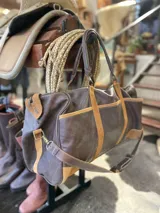 Bolso de viaje tipo duffel de cuero negro con detalles de refuerzo en gamuza y doble asa de mano.